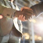 Low angle closeup shot of two business partners in handshake: unrecognizable African -American businessman shaking hands with Caucasian colleague in hall of modern glass office building at night time